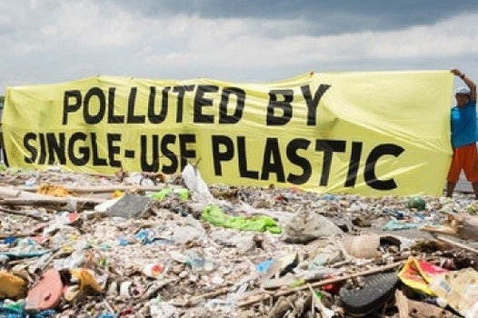 Polluted By Single-Use Plastic Sign on a shore behind beached plastic waste