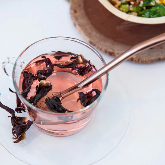 Glass tea cup containing herbal tea and a rose gold tea drinking spoon on a white surface, with a bowl of salad in the background.