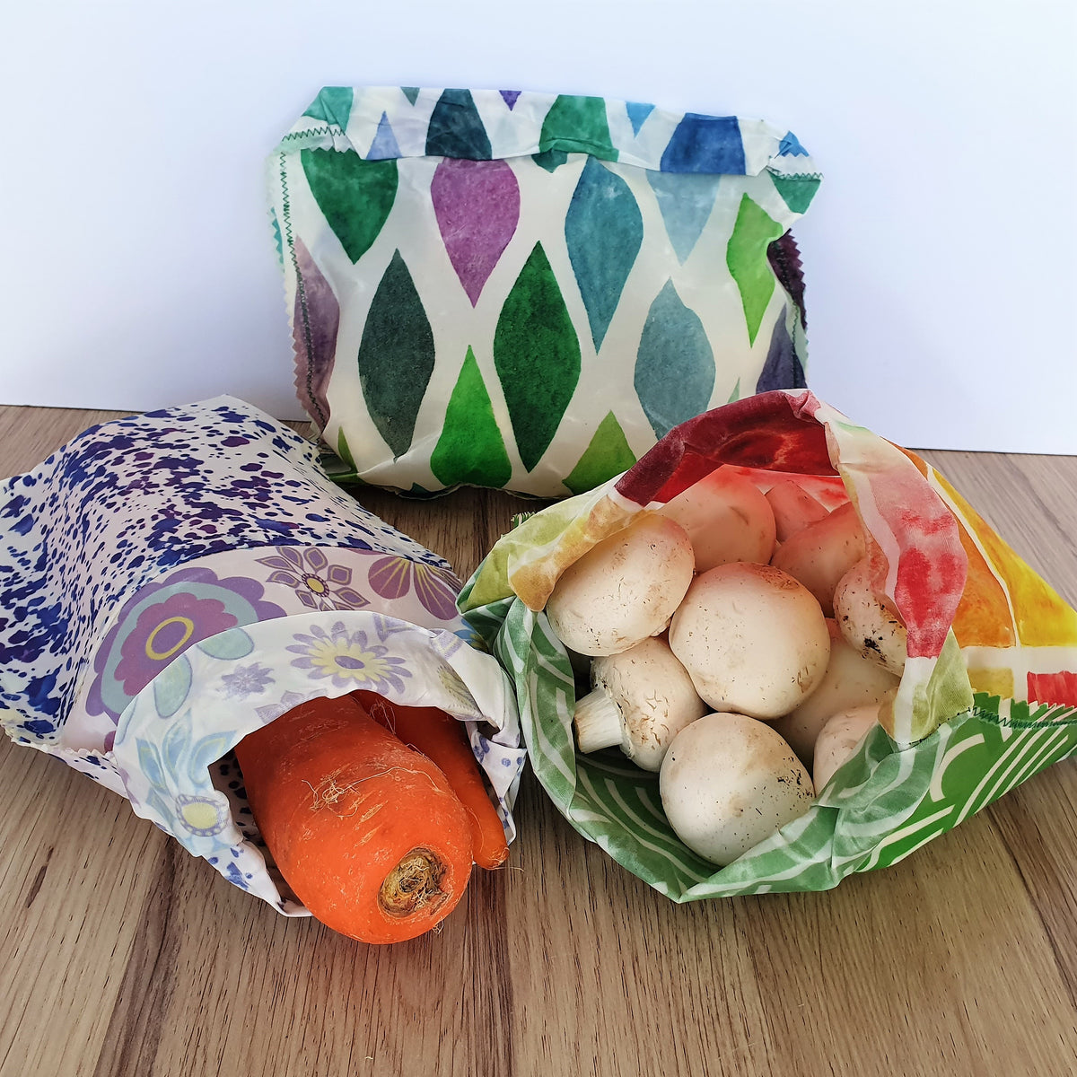 Three Reusable Beeswax Fabric Bags with Different Patterns Containing Vegetables on a Wooden Surface.