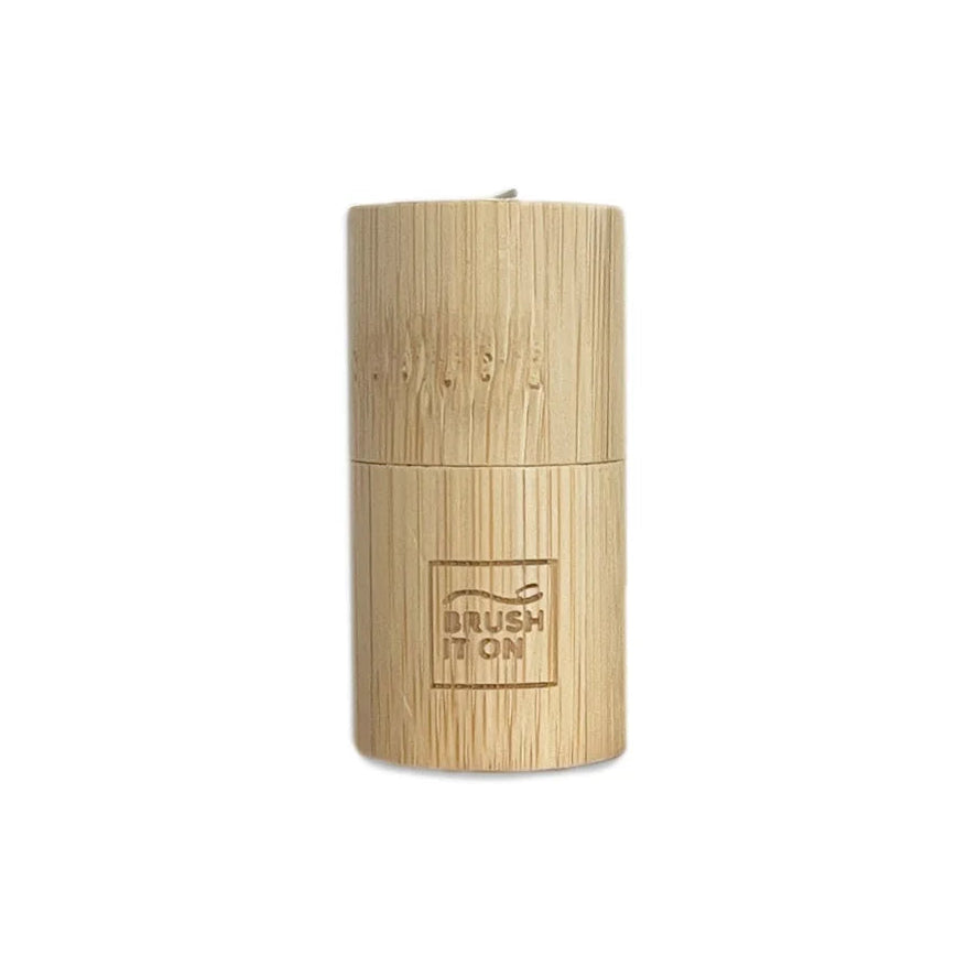 A cylindrical bamboo holder for corn starch floss, featuring a brushed metal brush head inserted at the top and an engraved logo that reads 'BRUSH IT ON'. 