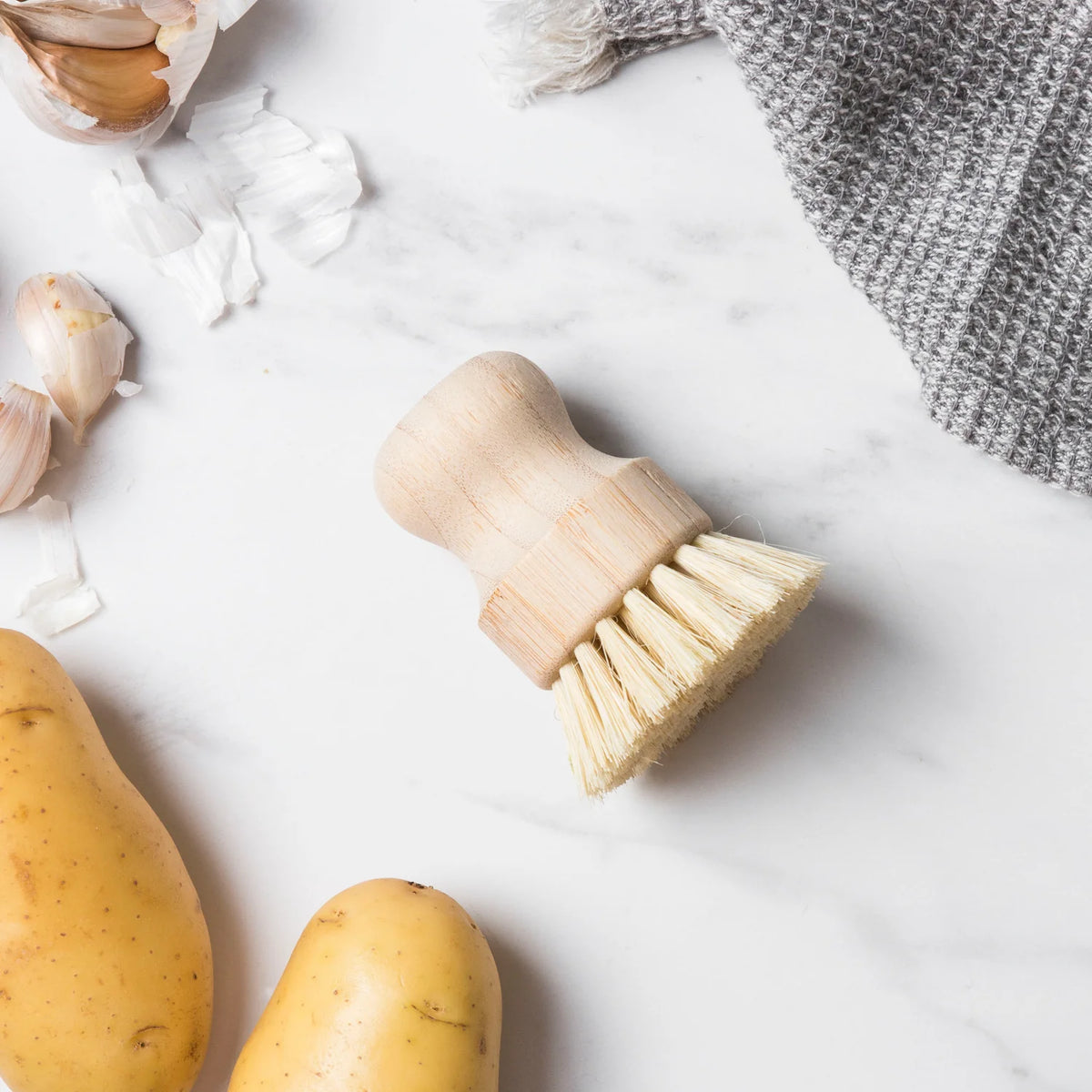 Wooden veggie scrubber on a marble surface with potatoes and garlic cloves.
