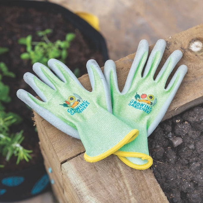 Green gardening gloves with yellow accents on a wooden tool next to a pot of plants.
