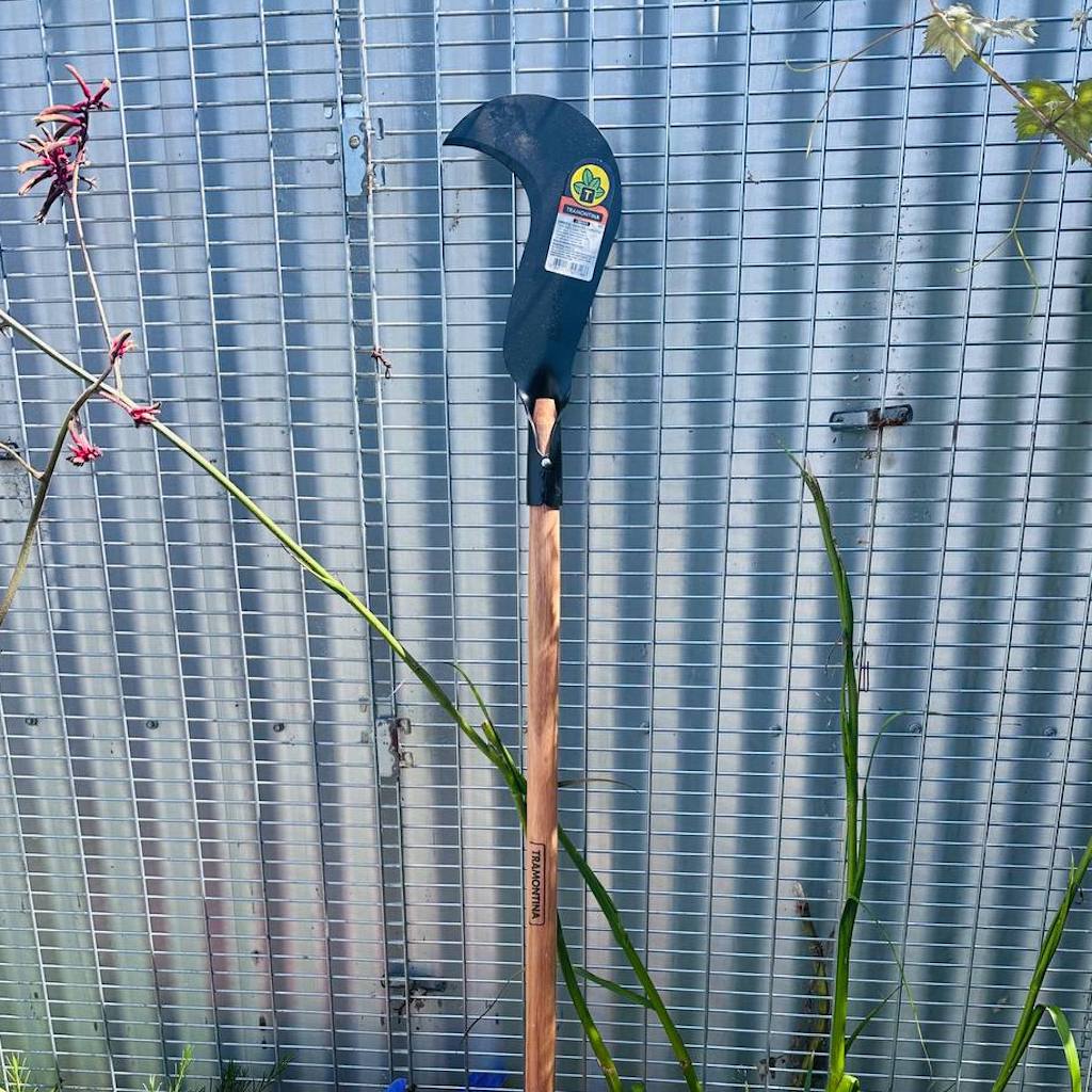 Carbon steel sickle with bill hook shape and long, hardwood handle leaning against a fence.