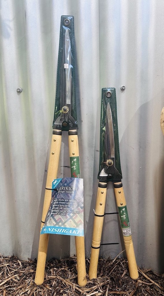 Long handled hedge shears displayed next to short handled hedge shears leaning on a wall.