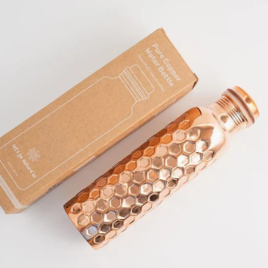 A pure copper water bottle with a diamond hammered pattern and a matching screw cap, shown next to its brown cardboard packaging box.