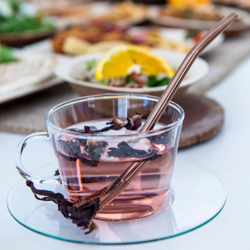 Clear glass cup containing tea ingredients and a rose gold tea drinking straw with a blurred background of a dining table with food.