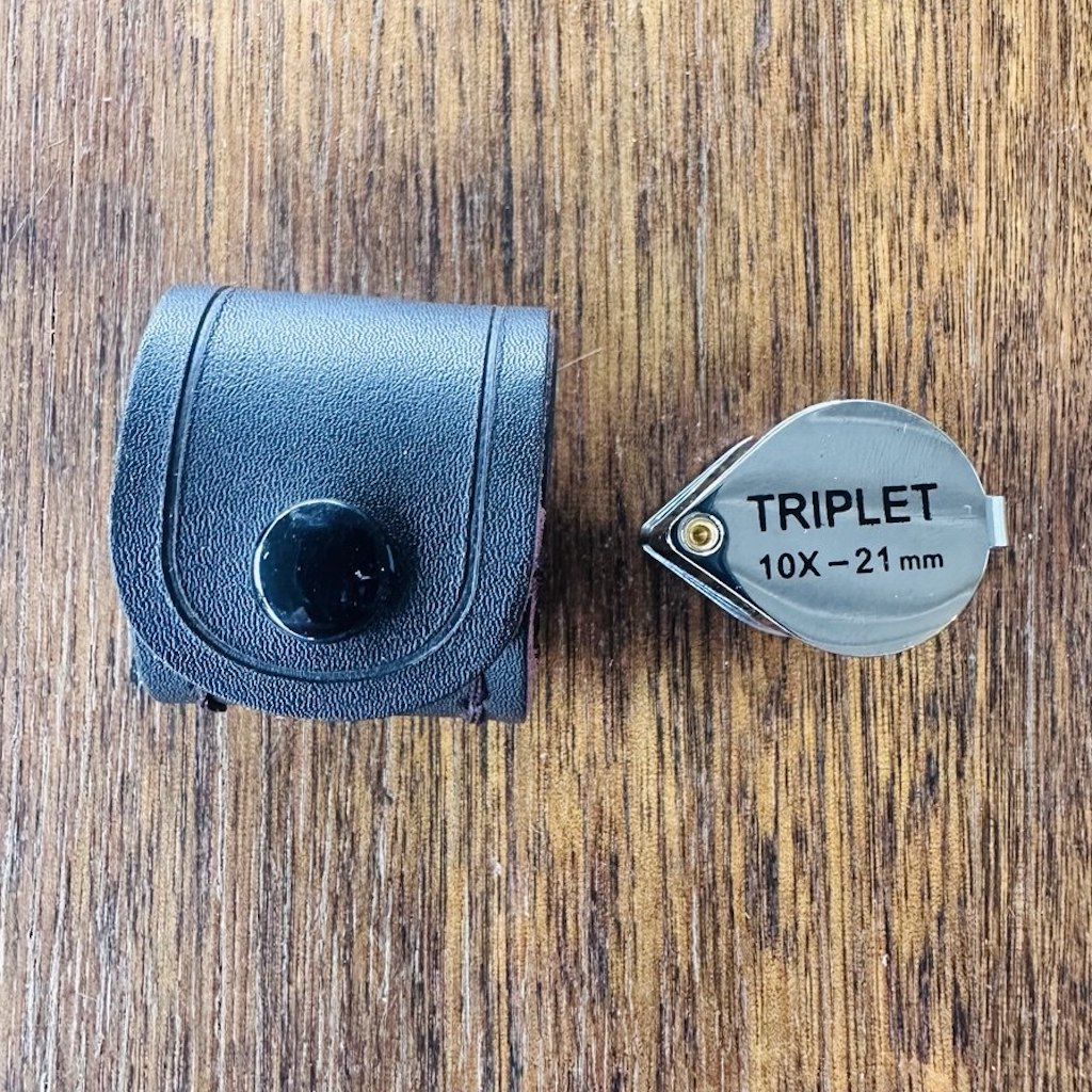 A compact hand held magnifying lens with words Triplet 10x – 21mm engraved on metal cover, pictured next to a black storage case.